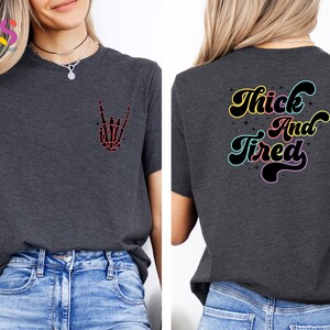 Op de afbeelding: Een donkergrijs T-shirt met een skelet hand die een rock-and-roll-teken maakt op de voorkant en de tekst "Thick and Tired" in regenboogkleuren op de achterkant.