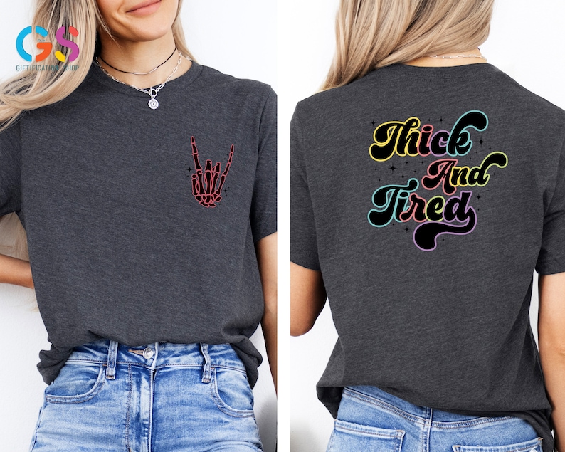 Op de afbeelding: Een donkergrijs T-shirt met een skelet hand die een rock-and-roll-teken maakt op de voorkant en de tekst "Thick and Tired" in regenboogkleuren op de achterkant.