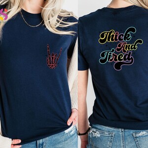 Op de afbeelding: Een marineblauw t-shirt met een skelethand die een rock-and-roll-teken maakt op de voorkant en de tekst "Thick And Tired" in regenboogkleuren op de achterkant.