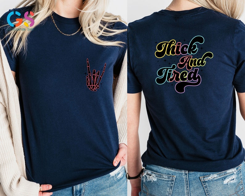 Op de afbeelding: Een marineblauw t-shirt met een skelethand die een rock-and-roll-teken maakt op de voorkant en de tekst "Thick And Tired" in regenboogkleuren op de achterkant.