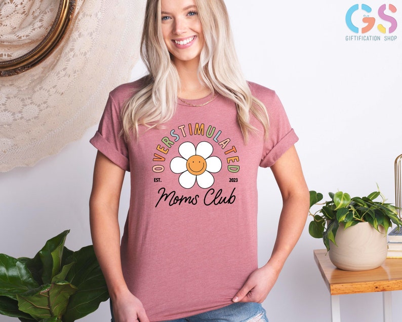 May include: A pink t-shirt with a white daisy and the text "Overstimulated Moms Club Est. 2023" in rainbow colors.