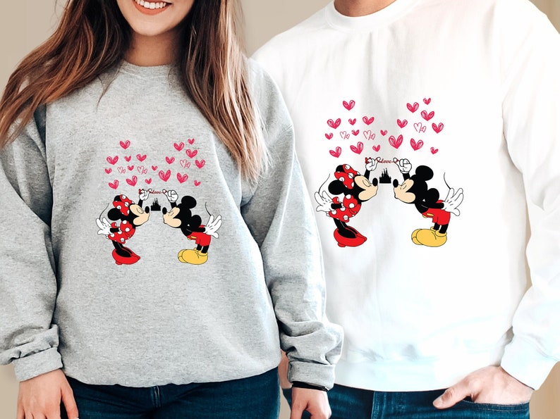 May include: Two sweatshirts, one gray and one white, feature a cartoon design of Mickey and Minnie Mouse kissing, with pink hearts floating above them. The design includes the word "Believe" and a castle silhouette.