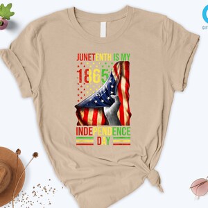 Puede incluir: Camiseta color beige con un dise&ntilde;o que celebra el Juneteenth. El dise&ntilde;o presenta una bandera estadounidense y el texto "Juneteenth is my 1865 Independence Day" en rojo, amarillo, verde y negro.