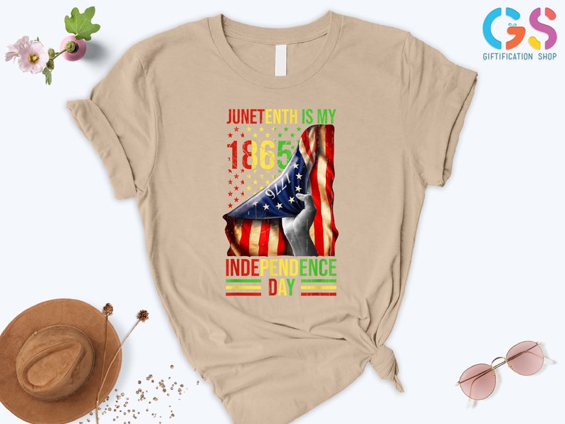 Puede incluir: Camiseta color beige con un dise&ntilde;o que celebra el Juneteenth. El dise&ntilde;o presenta una bandera estadounidense y el texto "Juneteenth is my 1865 Independence Day" en rojo, amarillo, verde y negro.