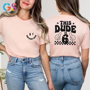 May include: Two pink t-shirts with black text. The front shirt has a smiley face with a lightning bolt and the text "Birthday Dude". The back shirt has the text "This Dude is 6" with two thumbs up and a checkered pattern.