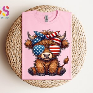 Puede incluir: Camiseta rosa con una vaca de las Tierras Altas de dibujos animados que lleva gafas de sol y una bandana, ambas decoradas con dise&ntilde;os de la bandera estadounidense. La vaca tiene pelaje marr&oacute;n y un aro en la nariz. La camiseta se muestra sobre una superficie tejida.