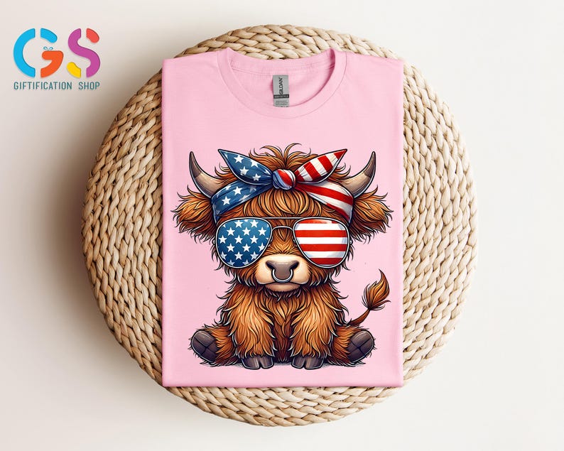 Puede incluir: Camiseta rosa con una vaca de las Tierras Altas de dibujos animados que lleva gafas de sol y una bandana, ambas decoradas con dise&ntilde;os de la bandera estadounidense. La vaca tiene pelaje marr&oacute;n y un aro en la nariz. La camiseta se muestra sobre una superficie tejida.
