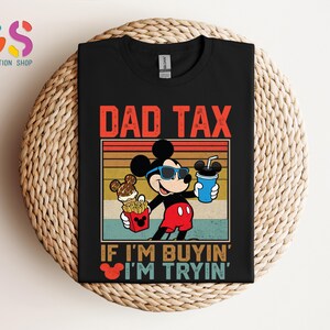 May include: Black t-shirt featuring Mickey Mouse wearing sunglasses and holding a drink and fries. The text "DAD TAX" is above the image, with "IF I'M BUYIN' I'M TRYIN'" below. The design has a retro, vintage style.