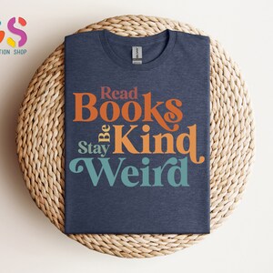 K&ouml;nnte beinhalten: Ein marineblaues T-Shirt mit einer Retro-Grafik, auf der "Read Books Be Kind Stay Weird" steht. Der Text ist in einem Regenbogen-Farbschema gehalten.