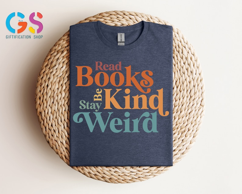 K&ouml;nnte beinhalten: Ein marineblaues T-Shirt mit einer Retro-Grafik, auf der "Read Books Be Kind Stay Weird" steht. Der Text ist in einem Regenbogen-Farbschema gehalten.