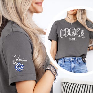 May include: A gray t-shirt with the text "CHEER MOM" in white on the front. The shirt has a blue and white pom-pom on the left sleeve with the name "Jessica" written above it.