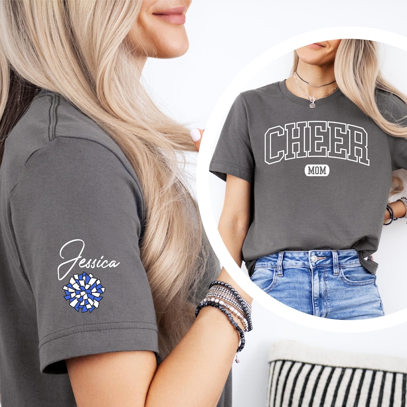 May include: A gray t-shirt with the text "CHEER MOM" in white on the front. The shirt has a blue and white pom-pom on the left sleeve with the name "Jessica" written above it.