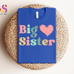 May include: A royal blue t-shirt with the text "Big Sister" in colorful retro font and a pink heart.