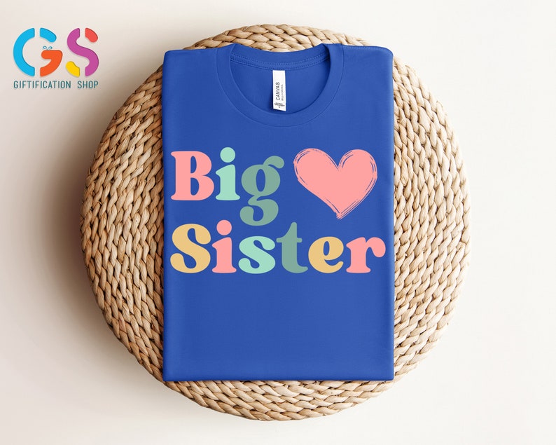 May include: A royal blue t-shirt with the text "Big Sister" in colorful retro font and a pink heart.