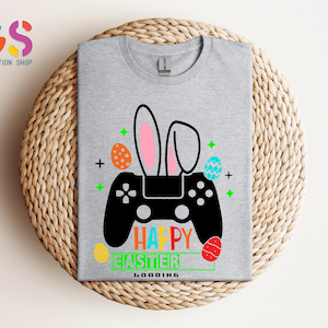 May include: A light gray t-shirt featuring a black controller design with bunny ears and colorful Easter eggs. The words "Happy Easter" and "Loading" are printed in green and yellow. The shirt is folded on a woven surface.