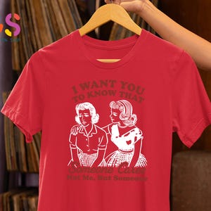 Op de afbeelding: Rood T-shirt met een witte afbeelding van twee vrouwen. De tekst op het shirt luidt "Ik wil dat je weet dat iemand geeft. Niet ik, maar iemand."