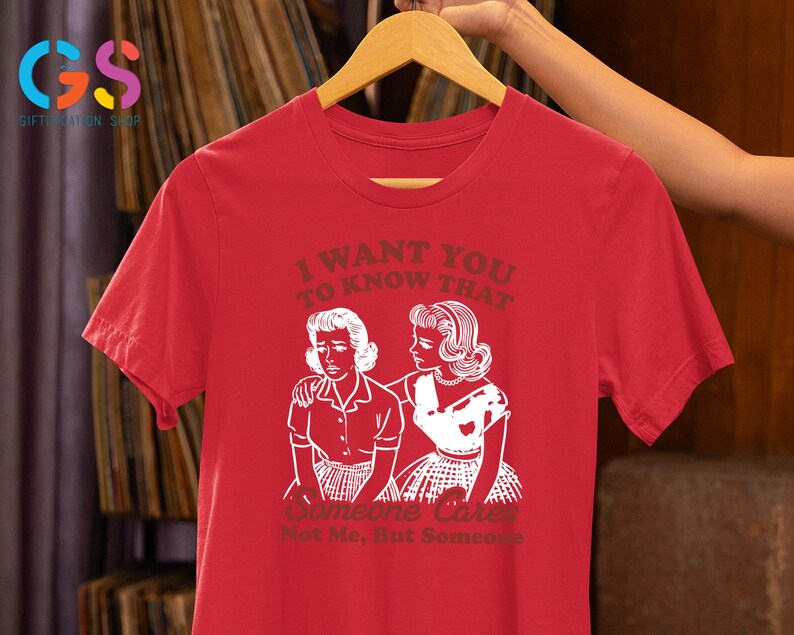 Op de afbeelding: Rood T-shirt met een witte afbeelding van twee vrouwen. De tekst op het shirt luidt "Ik wil dat je weet dat iemand geeft. Niet ik, maar iemand."