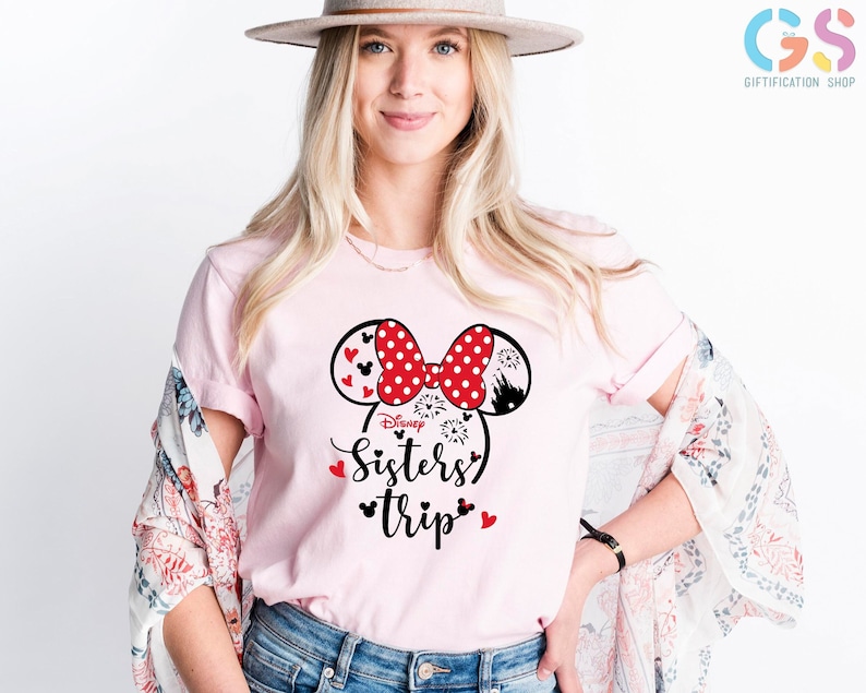 Puede incluir: Una camiseta rosa con un dise&ntilde;o de orejas de Mickey Mouse en blanco y negro. Las orejas tienen lunares rojos y un lazo rojo. El texto "Disney Sisters' Trip" est&aacute; impreso debajo de las orejas.