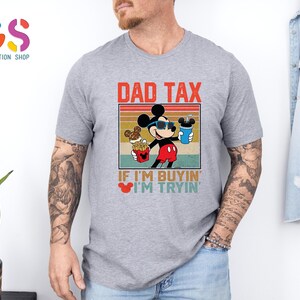 May include: A gray t-shirt featuring a graphic of Mickey Mouse wearing sunglasses and holding food and a drink. The text "DAD TAX" and "IF I'M BUYIN' I'M TRYIN'" are printed above and below the graphic, respectively.