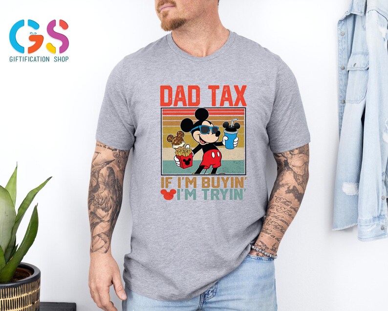 May include: A gray t-shirt featuring a graphic of Mickey Mouse wearing sunglasses and holding food and a drink. The text "DAD TAX" and "IF I'M BUYIN' I'M TRYIN'" are printed above and below the graphic, respectively.