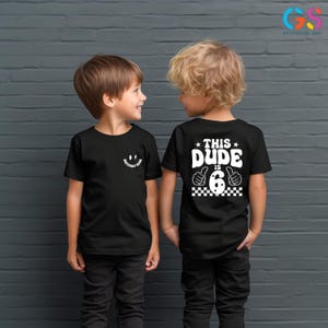 May include: Two children wearing black t-shirts. The child on the left has a smiley face with the text "Birthday Dude" on the front of their shirt. The child on the right has the text "This Dude is 6" with a thumbs up and a checkered pattern on the back of their shirt.