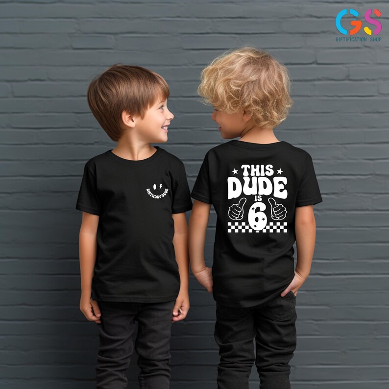 May include: Two children wearing black t-shirts. The child on the left has a smiley face with the text "Birthday Dude" on the front of their shirt. The child on the right has the text "This Dude is 6" with a thumbs up and a checkered pattern on the back of their shirt.
