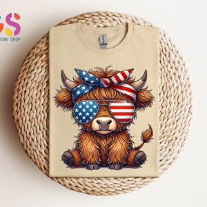 Puede incluir: Una camiseta beige con un dibujo animado de una vaca de las Tierras Altas que lleva gafas de sol y una diadema, ambas decoradas con dise&ntilde;os de la bandera americana. La vaca tiene pelaje marr&oacute;n y est&aacute; sentada. La camiseta est&aacute; sobre una superficie circular tejida.