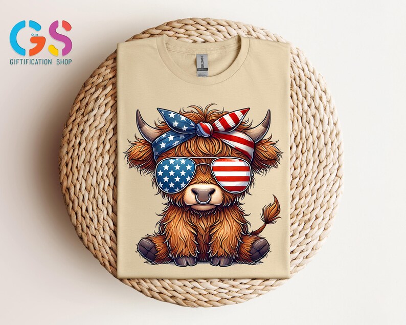Puede incluir: Una camiseta beige con un dibujo animado de una vaca de las Tierras Altas que lleva gafas de sol y una diadema, ambas decoradas con dise&ntilde;os de la bandera americana. La vaca tiene pelaje marr&oacute;n y est&aacute; sentada. La camiseta est&aacute; sobre una superficie circular tejida.