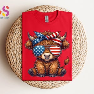 Puede incluir: Camiseta roja con una vaca de las Tierras Altas de dibujos animados que lleva gafas de sol y una diadema con un dise&ntilde;o de la bandera americana. La vaca tiene pelaje marr&oacute;n y un aro en la nariz. La camiseta se muestra sobre una superficie tejida.
