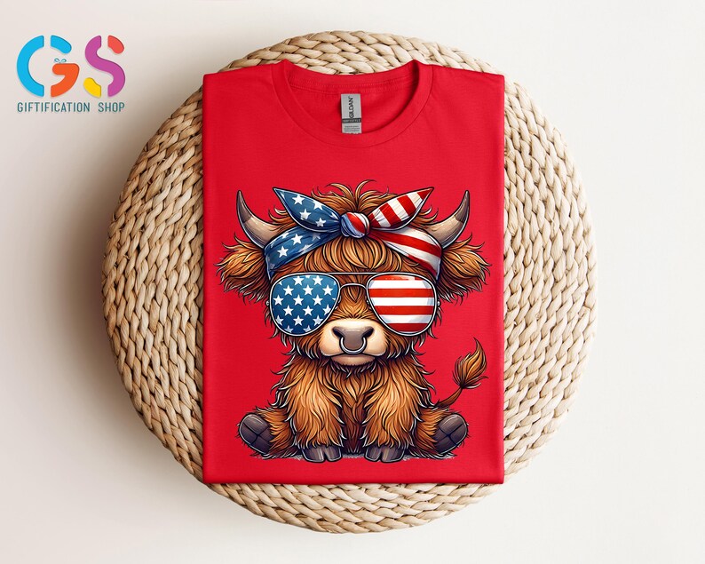 Puede incluir: Camiseta roja con una vaca de las Tierras Altas de dibujos animados que lleva gafas de sol y una diadema con un dise&ntilde;o de la bandera americana. La vaca tiene pelaje marr&oacute;n y un aro en la nariz. La camiseta se muestra sobre una superficie tejida.