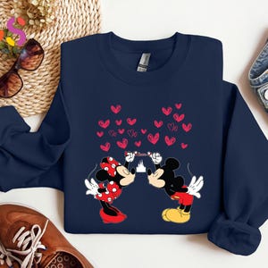 May include: Navy blue sweatshirt featuring Mickey and Minnie Mouse kissing, with a heart-shaped design. Minnie wears a red polka-dot bow and dress. Mickey wears red shorts and yellow shoes. A castle silhouette is between them.