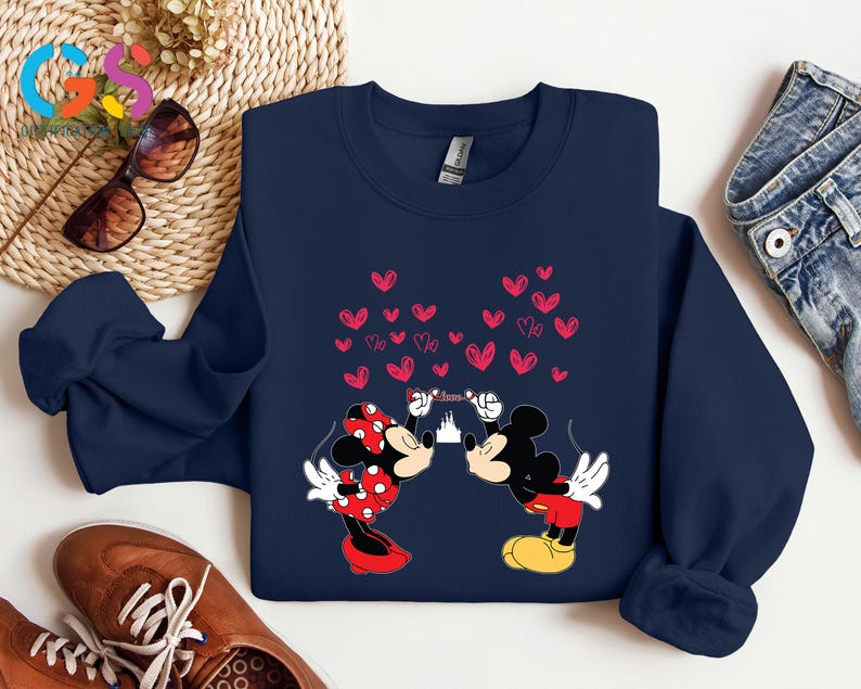 May include: Navy blue sweatshirt featuring Mickey and Minnie Mouse kissing, with a heart-shaped design. Minnie wears a red polka-dot bow and dress. Mickey wears red shorts and yellow shoes. A castle silhouette is between them.