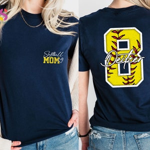 May include: Two navy blue t-shirts. The front of one shirt says "Softball Mom" with a heart. The back of the other shirt has a yellow softball with red stitching and the number 8 with the name "Deitzer" written in white.