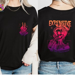 May include: Black t-shirt with a pink and orange gradient design. The front of the shirt features a skeleton hand making a peace sign with roses. The back of the shirt features a skeleton woman with a skull head drinking from a cup. The text "Expensive and Talks Back" is printed below the image.