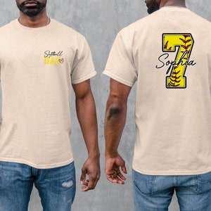 May include: A beige t-shirt with the text "Softball Dad" and a small heart on the front. The back of the shirt has a yellow softball with the number 7 and the name "Sophia" written in cursive.