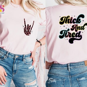Op de afbeelding: Roze T-shirt met een skelethand-afbeelding aan de voorkant en de tekst "Thick And Tired" op de achterkant. Het shirt wordt gedragen door iemand die blauwe jeans draagt.