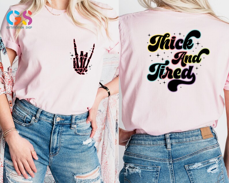 Op de afbeelding: Roze T-shirt met een skelethand-afbeelding aan de voorkant en de tekst "Thick And Tired" op de achterkant. Het shirt wordt gedragen door iemand die blauwe jeans draagt.