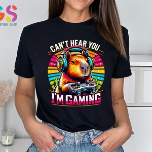 May include: Black t-shirt featuring a colorful graphic of a capybara wearing headphones and holding a game controller. Text on the shirt reads "CAN'T HEAR YOU I'M GAMING". The shirt has a crew neck and short sleeves.