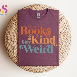 K&ouml;nnte beinhalten: Ein maroonfarbenes T-Shirt mit dem Text "Read Books Be Kind Stay Weird" in einer Retro-Schriftart. Die W&ouml;rter sind in verschiedenen Farben: orange, gelb und t&uuml;rkis.