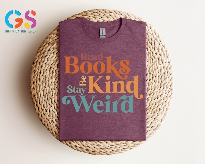 K&ouml;nnte beinhalten: Ein maroonfarbenes T-Shirt mit dem Text "Read Books Be Kind Stay Weird" in einer Retro-Schriftart. Die W&ouml;rter sind in verschiedenen Farben: orange, gelb und t&uuml;rkis.