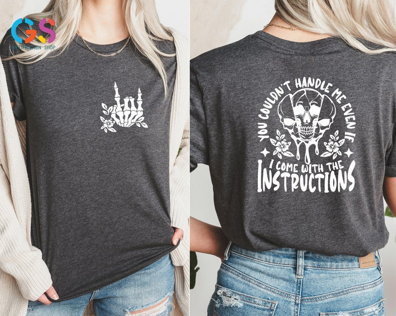 Op de afbeelding: Een donkergrijs T-shirt met een witte schedel en een bloemenontwerp op de achterkant. De tekst op de achterkant luidt "You couldn't handle me even if I come with the instructions". De voorkant van het shirt heeft een witte skelethand die het rock-and-roll-teken maakt met bloemmotieven.