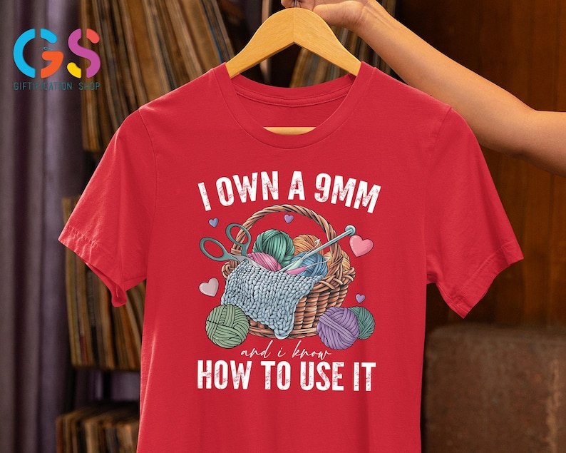 May include: Red t-shirt with a white graphic of a basket of yarn and knitting needles. The text "I own a 9mm and I know how to use it" is printed on the shirt.