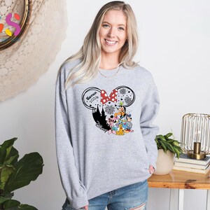 Pode incluir: Um moletom cinza claro com um gr&aacute;fico tem&aacute;tico da Disney. O gr&aacute;fico inclui uma silhueta da Minnie Mouse com um la&ccedil;o vermelho, o nome "Sophia 2024" e v&aacute;rios personagens da Disney. O design &eacute; definido contra uma silhueta preta de um castelo.