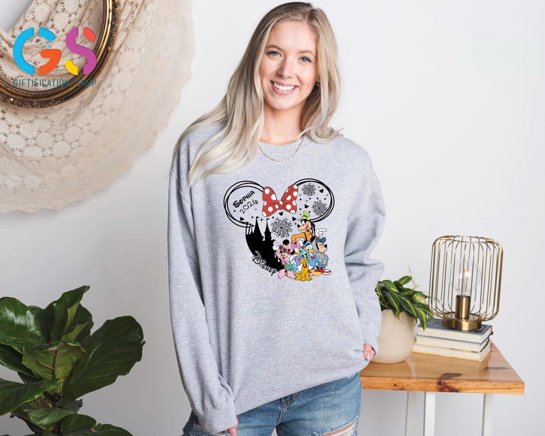 Pode incluir: Um moletom cinza claro com um gr&aacute;fico tem&aacute;tico da Disney. O gr&aacute;fico inclui uma silhueta da Minnie Mouse com um la&ccedil;o vermelho, o nome "Sophia 2024" e v&aacute;rios personagens da Disney. O design &eacute; definido contra uma silhueta preta de um castelo.