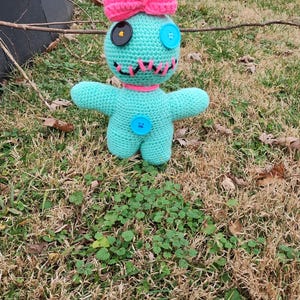 Op de afbeelding: Een handgemaakte turquoise gehaakte pop met een roze strik en knoopogen. De pop heeft een gestikte mond, een roze kraag en een blauwe knoop op de borst. De pop staat op een bed van gras.