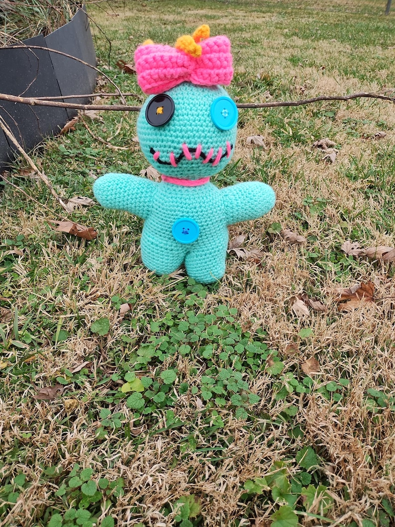 Op de afbeelding: Een handgemaakte turquoise gehaakte pop met een roze strik en knoopogen. De pop heeft een gestikte mond, een roze kraag en een blauwe knoop op de borst. De pop staat op een bed van gras.
