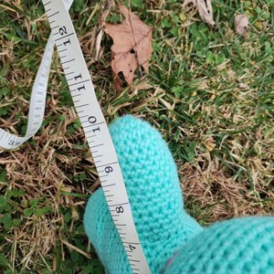 Op de afbeelding: Een close-up van een lichtblauwe gebreide want met een witte meetlint. De tape toont een meting van ongeveer 23 cm. De want ligt op groen gras.