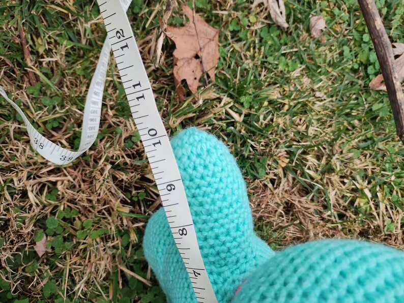 Op de afbeelding: Een close-up van een lichtblauwe gebreide want met een witte meetlint. De tape toont een meting van ongeveer 23 cm. De want ligt op groen gras.