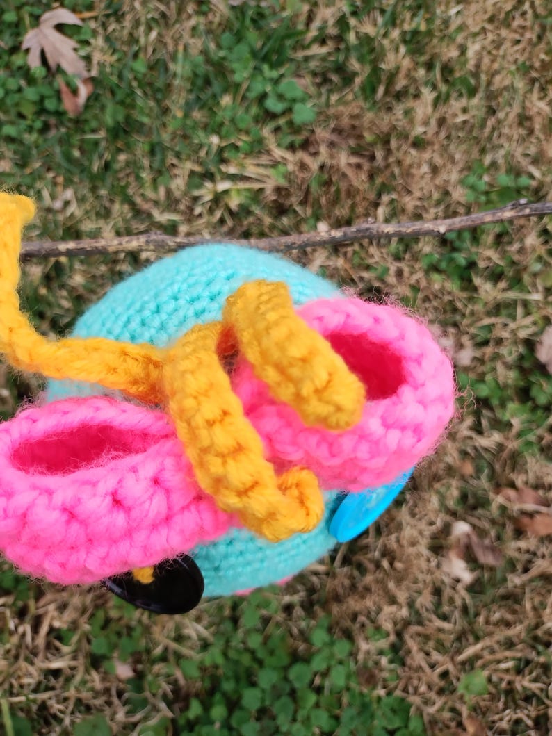 Op de afbeelding: Een handgemaakt, gehaakt speelgoed in de vorm van een kleurrijk voertuig. Het speelgoed heeft een lichtblauwe basis, roze wielen en gele accenten. Een blauwe knoop en zwart detail zijn ook zichtbaar. Het speelgoed staat tegen een achtergrond van groen gras en gevallen bladeren.