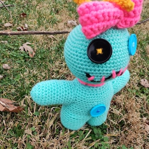 Op de afbeelding: Een gehaakte pop in turquoise met een roze strik, gele antennes en knoopogen. De pop heeft een gestikte roze mond en blauwe knoopaccenten. Het wordt buiten op gras gepresenteerd.